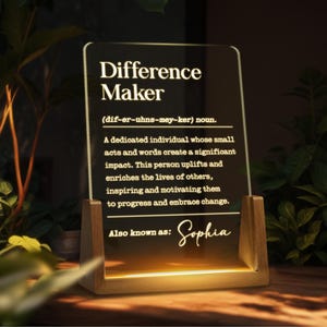 May include: A white acrylic sign with a wooden base, illuminated from below. The sign defines "Difference Maker" as a dedicated individual whose small acts and words create a significant impact. The sign also states "Also known as: Sophia".