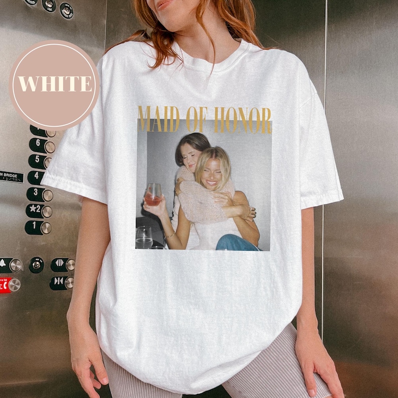 May include: White t-shirt with a gold "MAID OF HONOR" graphic and a photo of two women hugging. The shirt has a crew neck and short sleeves. A circular "WHITE" label is in the upper left corner. The shirt is a casual style.