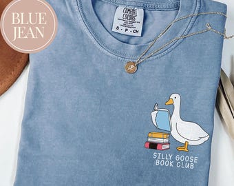 May include: A blue jean Comfort Colors t-shirt featuring a goose reading a book, with the text "Silly Goose Book Club". A gold necklace with a pendant is on the shirt.