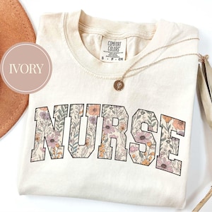May include: Ivory-colored t-shirt with the word "NURSE" in large, floral-patterned letters. The shirt has a crew neck and a small tag at the top. A gold necklace with a pendant is draped across the shirt.