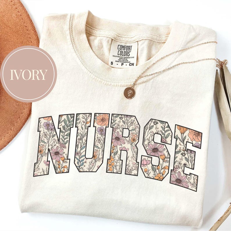 May include: Ivory-colored t-shirt with the word "NURSE" in large, floral-patterned letters. The shirt has a crew neck and a small tag at the top. A gold necklace with a pendant is draped across the shirt.