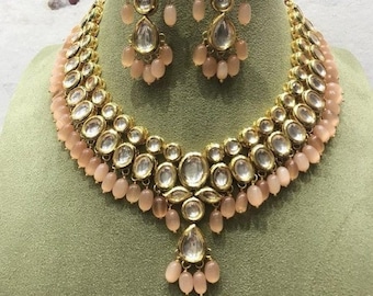 May include: A gold necklace and earrings set with a multi-layered design featuring large, clear, round stones and smaller, peach-colored beads.