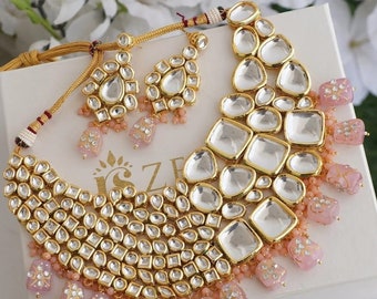May include: A gold and pink necklace set with large, clear, faceted stones and pink, floral-patterned beads. The necklace is layered with multiple rows of smaller stones. The earrings are a matching design with smaller stones and beads.
