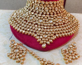 May include: A gold necklace set featuring a large, ornate choker, matching earrings, and a long, beaded maang tikka. The set is decorated with clear, faceted stones.