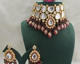 May include: A gold and silver necklace with a large, teardrop-shaped pendant and red and green beads. The necklace is on a green velvet stand. A pair of matching earrings are also shown.