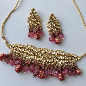 May include: A gold choker necklace with pink and white beads and a matching pair of earrings. The necklace features a large, ornate design with multiple layers of beads and a central piece of gold.
