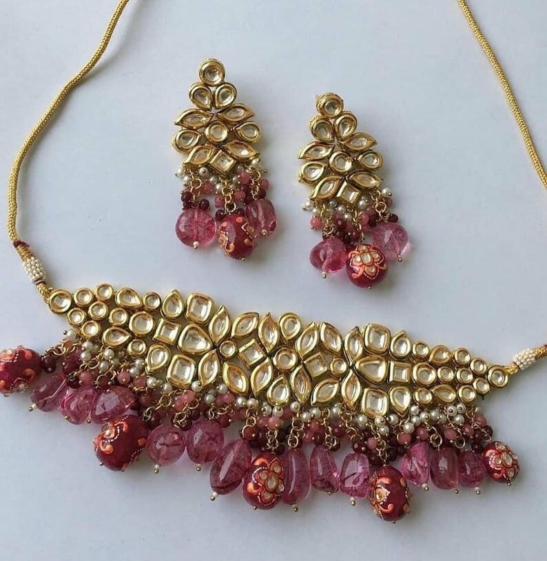 May include: A gold choker necklace with pink and white beads and a matching pair of earrings. The necklace features a large, ornate design with multiple layers of beads and a central piece of gold.
