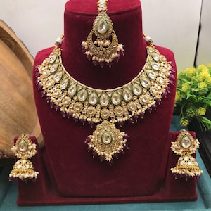 Op de afbeelding: Een gouden en witte Indiase bruids kettingset met groene en paarse accenten. De set bevat een ketting, oorbellen en een maang tikka.