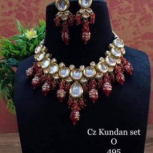 May include: A gold necklace and earrings set with red and clear stones. The necklace features a large, round pendant with a clear stone and smaller, red stones. The earrings are small and have a similar design to the necklace. The set is labelled "Cz Kundan set O 495".