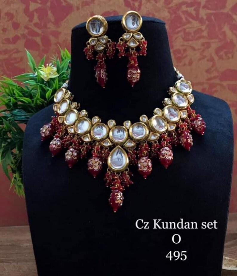 May include: A gold necklace and earrings set with red and clear stones. The necklace features a large, round pendant with a clear stone and smaller, red stones. The earrings are small and have a similar design to the necklace. The set is labelled "Cz Kundan set O 495".