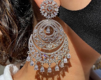 May include: A pair of large, silver earrings with a circular design. The earrings are adorned with many small, clear stones that create a sparkling effect.