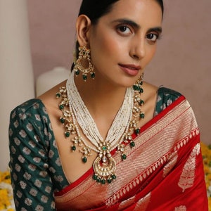 May include: A woman wearing a red and gold saree with a green blouse. She is wearing a pearl and emerald necklace and matching earrings.