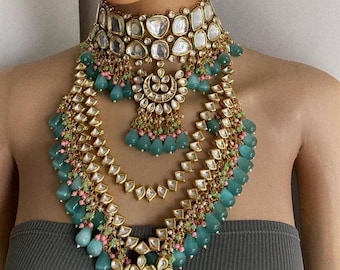 May include: A multi-layered gold necklace with teardrop-shaped green and pink stones. The necklace features a large, ornate choker with a floral design and a pendant with a crescent moon shape.