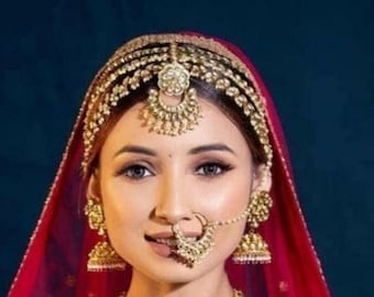 May include: A woman wearing a red veil and a gold headpiece with intricate designs. She is also wearing a gold nose ring and earrings.