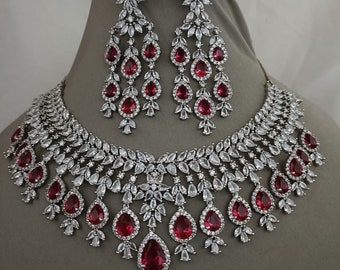 May include: A silver necklace and earring set featuring ruby-red, teardrop-shaped gemstones. The necklace and earrings are embellished with clear, sparkling accents. The set is displayed on a mannequin, highlighting the intricate design and craftsmanship.