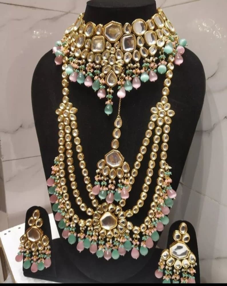 May include: A gold-tone Indian bridal necklace set with multiple strands, featuring large, mirrored, round and octagonal stones. The set includes a choker, a long necklace, and matching earrings with pink and green teardrop-shaped beads.