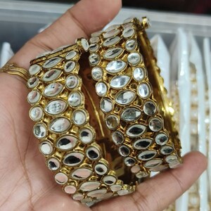 May include: Gold-coloured bangles adorned with clear, oval-shaped embellishments. The jewellery is held in a hand, highlighting the detailed design and reflective surfaces. The bangles appear to be part of a larger collection of similar items.