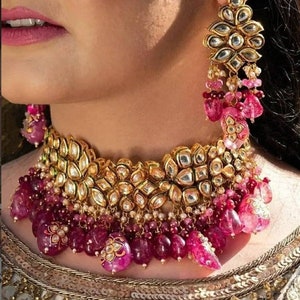 May include: A gold and pink choker necklace with a large, ornate design. The necklace is decorated with small, round stones and has a dangling pendant of pink stones.