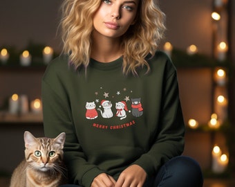 May include: An olive green sweatshirt featuring a festive design of four cartoon cats wearing holiday attire, with the text "MERRY CHRISTMAS" below. The cats are adorned with sweaters, hats, and scarves. The sweatshirt is worn by a person.