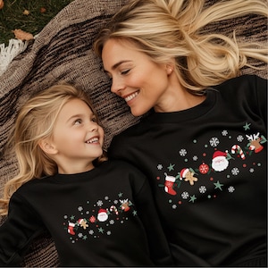 May include: Black sweatshirts with a Christmas design. The sweatshirts feature a Santa Claus, reindeer, candy cane, stocking, and gingerbread man design with snowflakes and stars. The sweatshirts are worn by a mother and daughter.