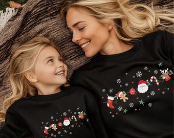 May include: Black sweatshirts with a Christmas design. The sweatshirts feature a Santa Claus, reindeer, candy cane, stocking, and gingerbread man design with snowflakes and stars. The sweatshirts are worn by a mother and daughter.