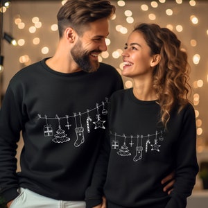 May include: Black sweatshirts with a white line drawing of Christmas ornaments, a tree, a stocking, and a candy cane. The sweatshirts are worn by a smiling couple, with a blurred background of warm lights.