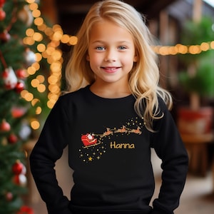 May include: A black long-sleeve sweatshirt featuring a graphic print of Santa Claus in a red sleigh pulled by three reindeer, surrounded by gold stars and the name Hanna printed in a yellow script font across the centre of the chest.