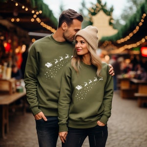 May include: Two people wearing olive green sweatshirts with a white graphic of Santa's sleigh and reindeer ascending with stars. The sweatshirts are long-sleeved and crew neck. The background is a blurred outdoor scene with string lights.