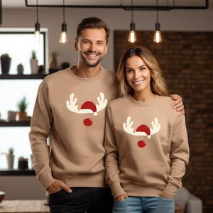 May include: Two people wearing matching tan sweatshirts with a reindeer face design. The design includes white antlers, a red nose, and a red Santa hat. The sweatshirts are a festive holiday theme.