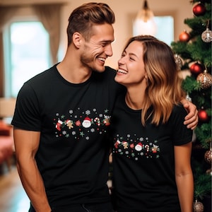 May include: A couple wearing matching black t-shirts with a Christmas-themed design featuring Santa Claus, a gingerbread man, a stocking, candy canes, and a reindeer. The design is repeated on both shirts.
