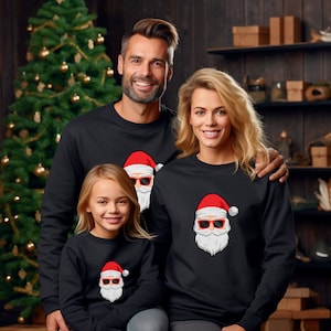 May include: Three people wearing matching black sweatshirts with a Santa Claus graphic. The Santa design features a red hat, sunglasses, and a white beard. A Christmas tree is visible in the background, suggesting a holiday theme.