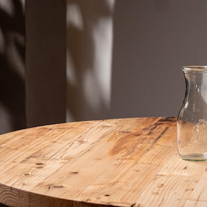 Könnte beinhalten: Eine klare Milchflasche aus Glas mit schmalem Hals und breitem Boden steht auf einem runden, hellen Holztisch. Die Flasche hat ein dekoratives Emblem auf der Vorderseite. Der Tisch hat eine natürliche Holzmaserung.