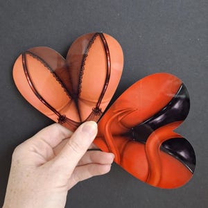 Può includere: Due adesivi a forma di cuore. Uno presenta un design di lingerie con finiture in pizzo e un colore marrone rossiccio. L'altro adesivo ha un design rosso e nero a tema diabolico. Gli adesivi sono tenuti su uno sfondo scuro.
