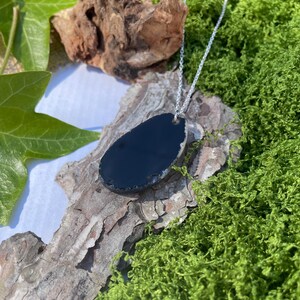 May include: A black teardrop-shaped pendant necklace with a silver chain. The pendant has a rough, natural edge and is set against a backdrop of green moss, wood, and leaves. The necklace is a simple, elegant accessory.