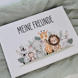 Könnte beinhalten: Ein weißer Buchumschlag mit dem Text "MEINE FREUNDE" und Illustrationen von Zeichentieren, darunter ein Zebra, ein Affe, ein Elefant, eine Giraffe, ein Löwe und ein Nilpferd. Die Tiere sind von grünen Blättern und braunem Laub umgeben.