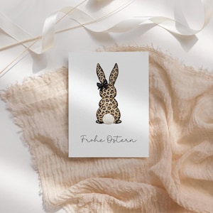 Könnte beinhalten: Eine Grußkarte mit einer Aquarellillustration eines Hasen mit Leopardenmuster. Der Hase hat eine schwarze Schleife und einen flauschigen weißen Schwanz. Die Worte "Frohe Ostern" sind darunter in Schreibschrift geschrieben.
