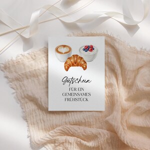 Könnte beinhalten: Eine weiße Karte mit Aquarellillustrationen eines Croissants, eines Cappuccinos mit herzförmigem Schaum und einer Schüssel Joghurt mit Beeren. Die Karte trägt den Text "Gutschein FÜR EIN GEMEINSAMES FRÜHSTÜCK" in schwarzer Schrift.