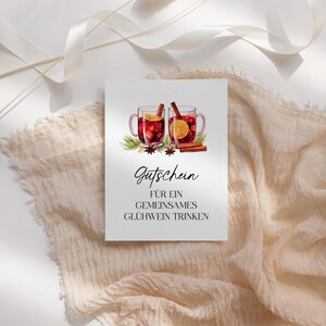 May include: A white card with a watercolor illustration of two mugs of mulled wine, cinnamon sticks, and orange slices. The card has the German word "Gutschein" and the phrase "FÜR EIN GEMEINSAMES GLÜHWEIN TRINKEN" printed on it.