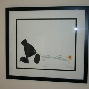 May include: Black and white print of a stylised bird pulling a flower with a yellow centre. The print is framed in a black frame and has the artist's signature "Nene" in the bottom right corner.