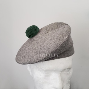 Puede incluir: Una boina de lana gris con un pompón verde oscuro. La boina se muestra en una cabeza de maniquí. La palabra "STITCHIFY" está impresa en la boina.