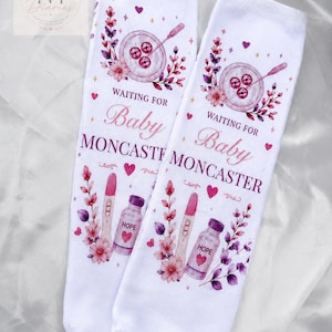 May include: A pair of white socks featuring a pink and purple floral design, a petri dish with three embryos, a pregnancy test, and a medicine bottle labelled HOPE. The text reads WAITING FOR Baby MONCASTER, accented with small hearts and stars.