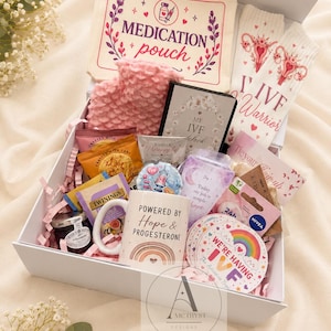 May include: A white gift box containing a cream medication pouch, white socks with uterus illustrations and IVF Warrior text, a pink textured hot water bottle, a mug with a rainbow and Powered by Hope & Progesterone text, and various snacks and small items.