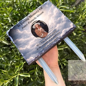 May include: A memorial plaque with the text "In Loving Memory Of Janet Pamela Green" and a photo. The plaque has a cloudy sky background and is held by two silver stakes. The text "May love hold you gently..." is also present.