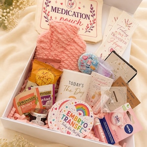 Può includere: Una scatola regalo bianca contenente una borsa dell'acqua calda rosa testurizzata, una pochette per medicinali color crema, una tazza bianca con la scritta Today's The Day, un sottobicchiere rotondo Embryo Transfer, bustine di tè Twinings, un balsamo labbra Nivea e vari piccoli oggetti.