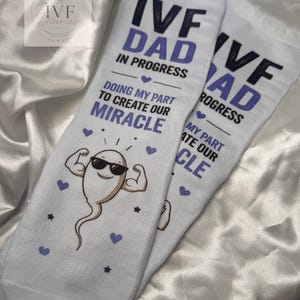May include: A pair of white crew socks featuring the text IVF DAD IN PROGRESS and DOING MY PART TO CREATE OUR MIRACLE in dark blue and purple lettering. The design includes a cartoon sperm character wearing sunglasses and flexing its arm muscles.
