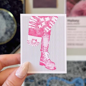 Könnte beinhalten: Ein rosa-weißer Aufkleber mit einer Grafik eines Beins, das Jeans und einen Stiefel trägt. Der Hintergrund ist ein weiß-schwarzes Streifenmuster.