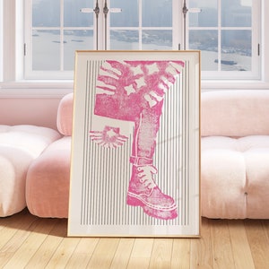 Könnte beinhalten: Ein gerahmter rosa-weißer Druck eines Beins, das einen Stiefel und Jeans trägt. Der Druck ist auf einem gestreiften Hintergrund. Der Rahmen ist golden.