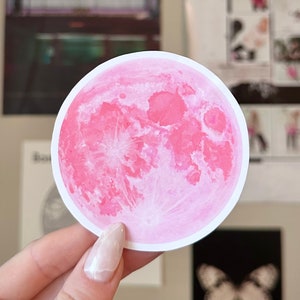 May include: A round sticker featuring a pink moon with a detailed surface. The moon is set against a white border. The sticker is held by a person.