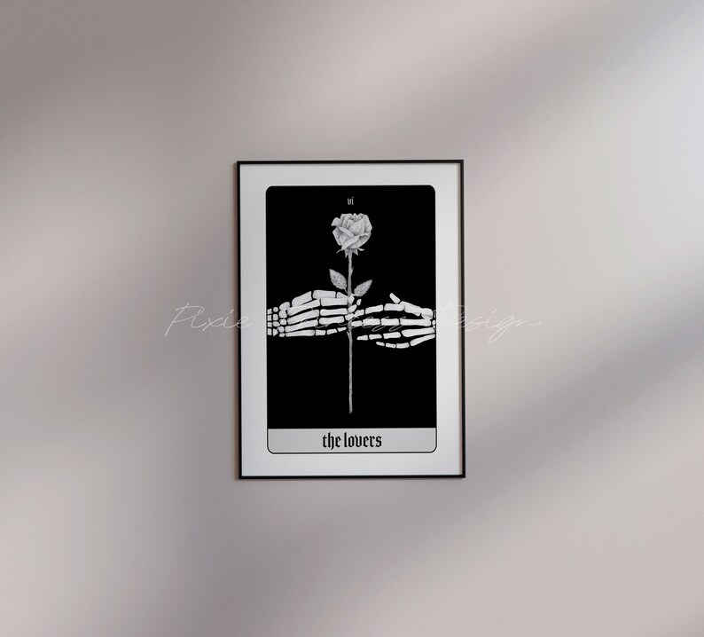 Puede incluir: Ilustraci&oacute;n de carta de tarot en blanco y negro de la carta de Los Enamorados. Dos manos esquel&eacute;ticas sostienen una sola rosa blanca con un tallo negro. La carta est&aacute; enmarcada en negro y tiene el texto "the lovers" en la parte inferior.