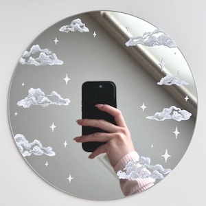 May include: A round mirror with a white painted design of clouds and stars. The mirror is reflecting a person holding a black smartphone.